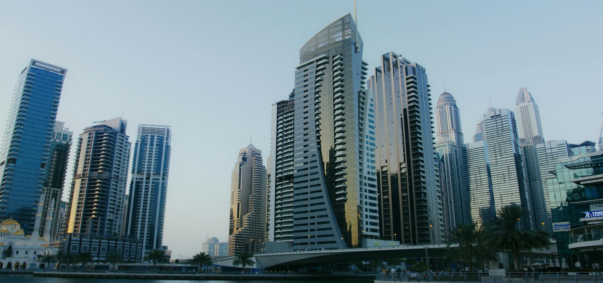 Skyline of modern skyscrapers under a clear blue sky, featuring diverse architecture and palm trees for greenery.