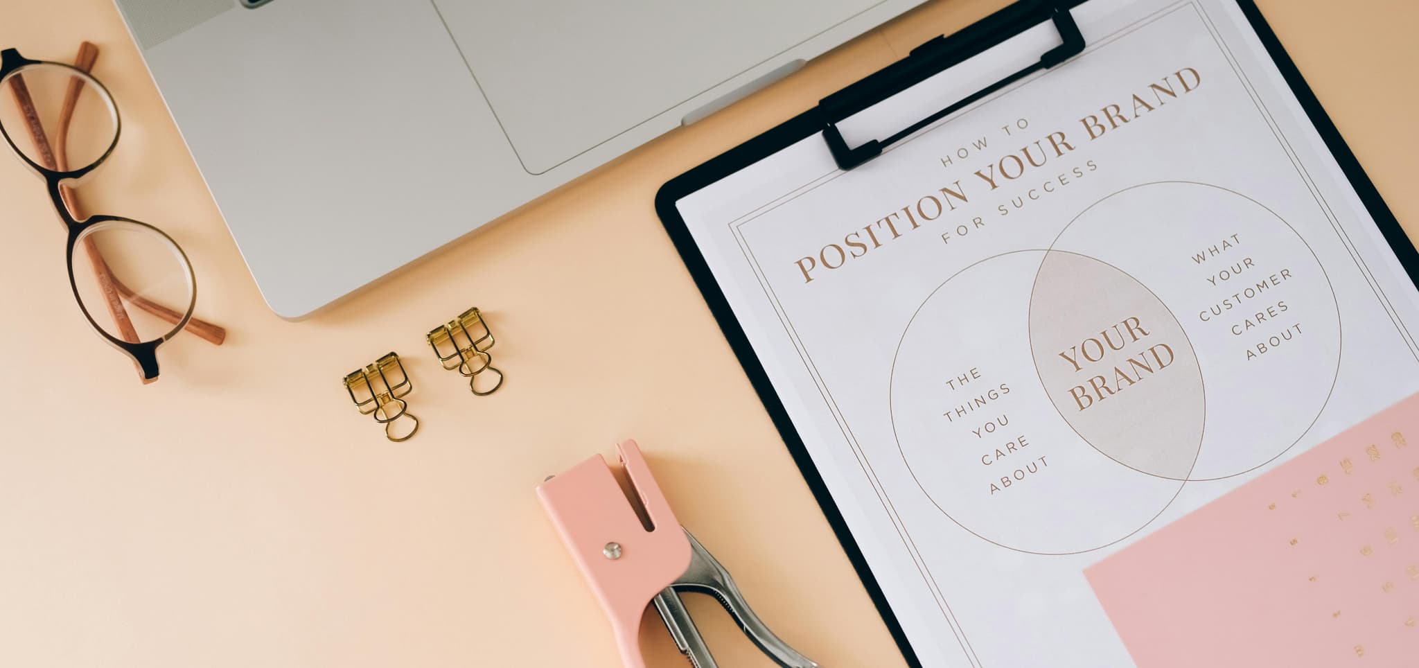 Flat lay of a laptop, eyeglasses, gold clips, pink stapler, and a clipboard with a branding diagram, exuding professionalism.